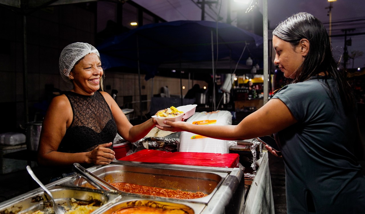 Mulher entregando um pratinho para outra numa barraca de comida